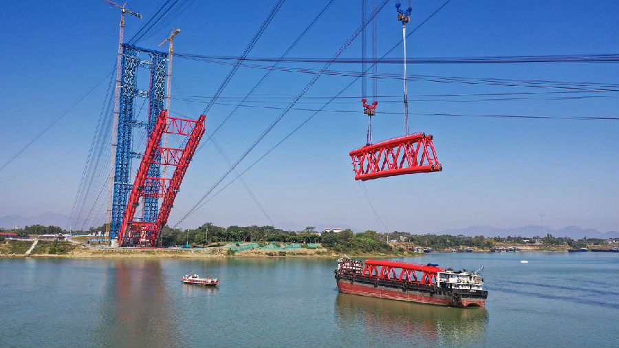 纜索吊裝系統由主索道系統和工作索道系統組成，其中主承載索為纜索吊裝系統最核心的受力結構，由16根密封鋼絲繩組成，單根長度2200米，如果將整個纜索吊裝系統使用到的鋼絲繩首尾相接，總長達到約99520米，相當于廣西南寧到欽州的直線距離。圖為拱肋運送到江面后，纜索吊裝系統進行吊裝。（梁暘善 攝）