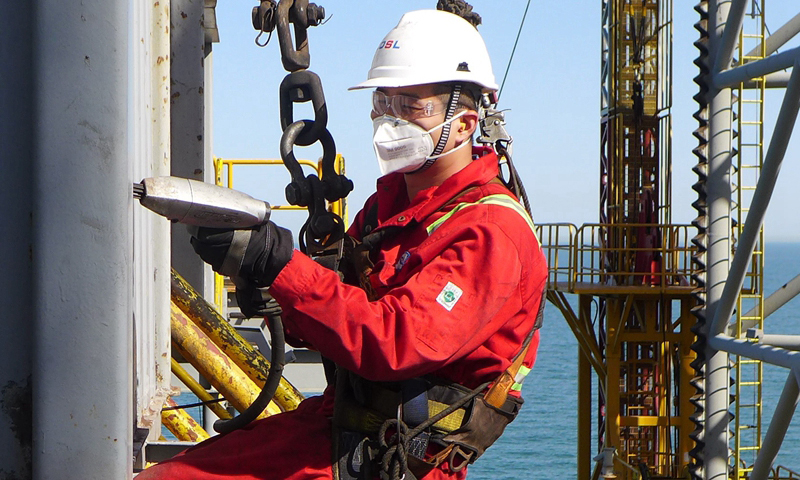 近日，海洋石油“932鉆井平臺”在渤中19-4油田進行三開鉆井作業。在平臺鉆進作業間隙，鉆井班組對平臺日常防腐作業難點區域展開防腐攻堅，徹底清除防腐工作死角，力保平臺安全。圖為平臺副司鉆黃鑫乘坐載人絞車對鉆臺擋風板外側進行除銹防腐作業。