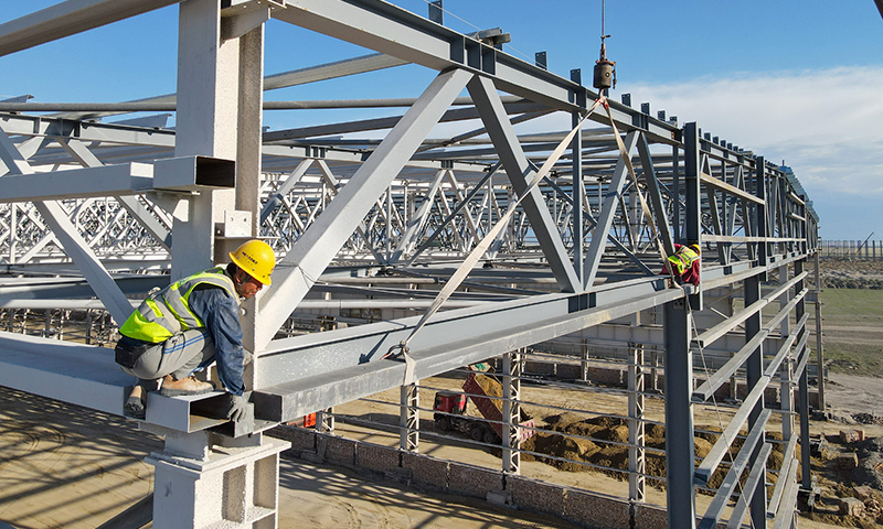 進入施工旺季，新疆塔城重點開發開放試驗區工程建設有序進行，來自中鐵十五局等多家單位的工人晝夜奮戰在建設現場。新疆塔城重點開發開放試驗區是我國西北地區第一個沿邊重點開發開放試驗區。圖為工人在20余米高的新疆塔城重點開發開放試驗區一處風機葉片制造項目頂部進行屋面檁條安裝作業（5月18日無人機拍攝）。新華社記者 高晗 攝